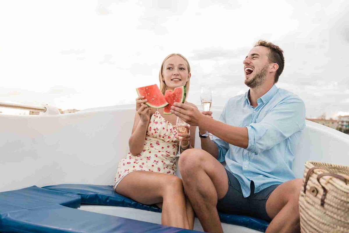 Happy young couple cheering with champagne and eating fruits on sail boat in summer vacation